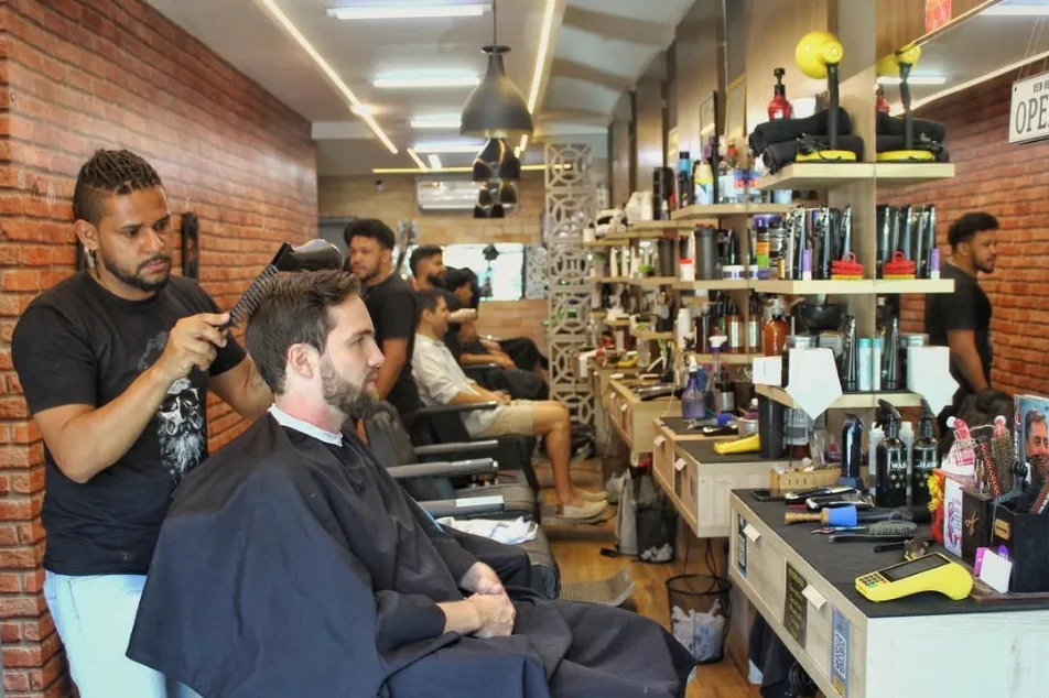 Salão da Antonio Barbearia em atendimento: um barbeiro escova o cabelo de um cliente na cadeira, com parede de tijolo aparente, luz linear no teto e a bancada de produtos ao fundo.