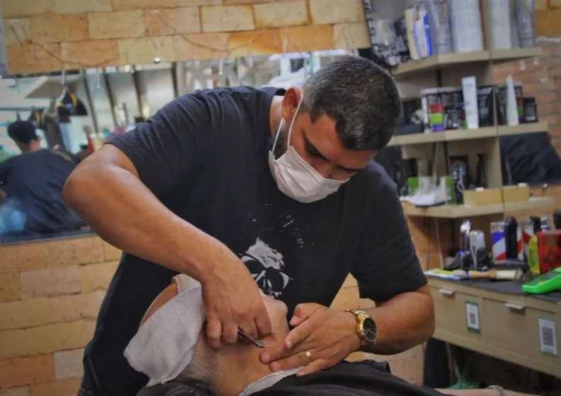 Barbeiro de máscara alinha a barba de um cliente reclinado na cadeira, com toalha sob o rosto.