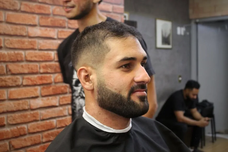 Cliente de barba aparada e degradê nas laterais, sentado com a capa preta diante da parede de tijolo.