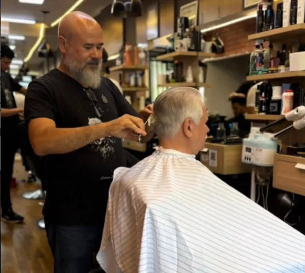 Barbeiro corta com tesoura o cabelo grisalho de um cliente, entre as prateleiras de produtos do salão.