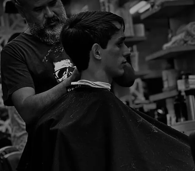 Fotografia em preto e branco: barbeiro finaliza o corte de um cliente na cadeira.