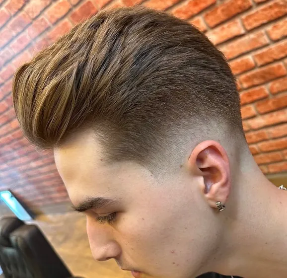 Topete alto penteado para trás, com degradê fechado nas laterais, diante da parede de tijolo.